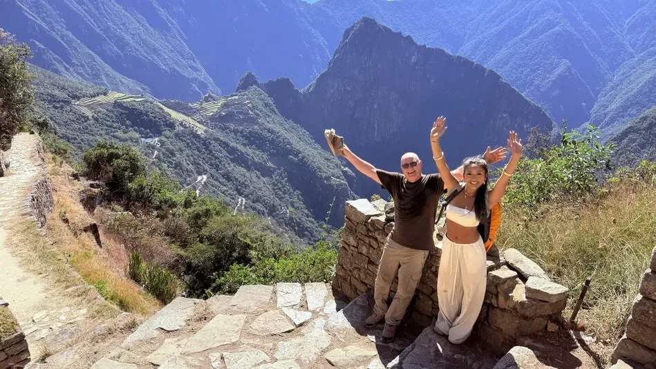 Two travelers celebrating at an Inca site with Machu Picchu and the Andes visible in the distance